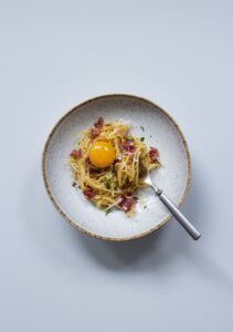 En grå skål med en tallrik med spagetti och vildsvinskött med en äggula på toppen. En gaffel ligger i tallriken.