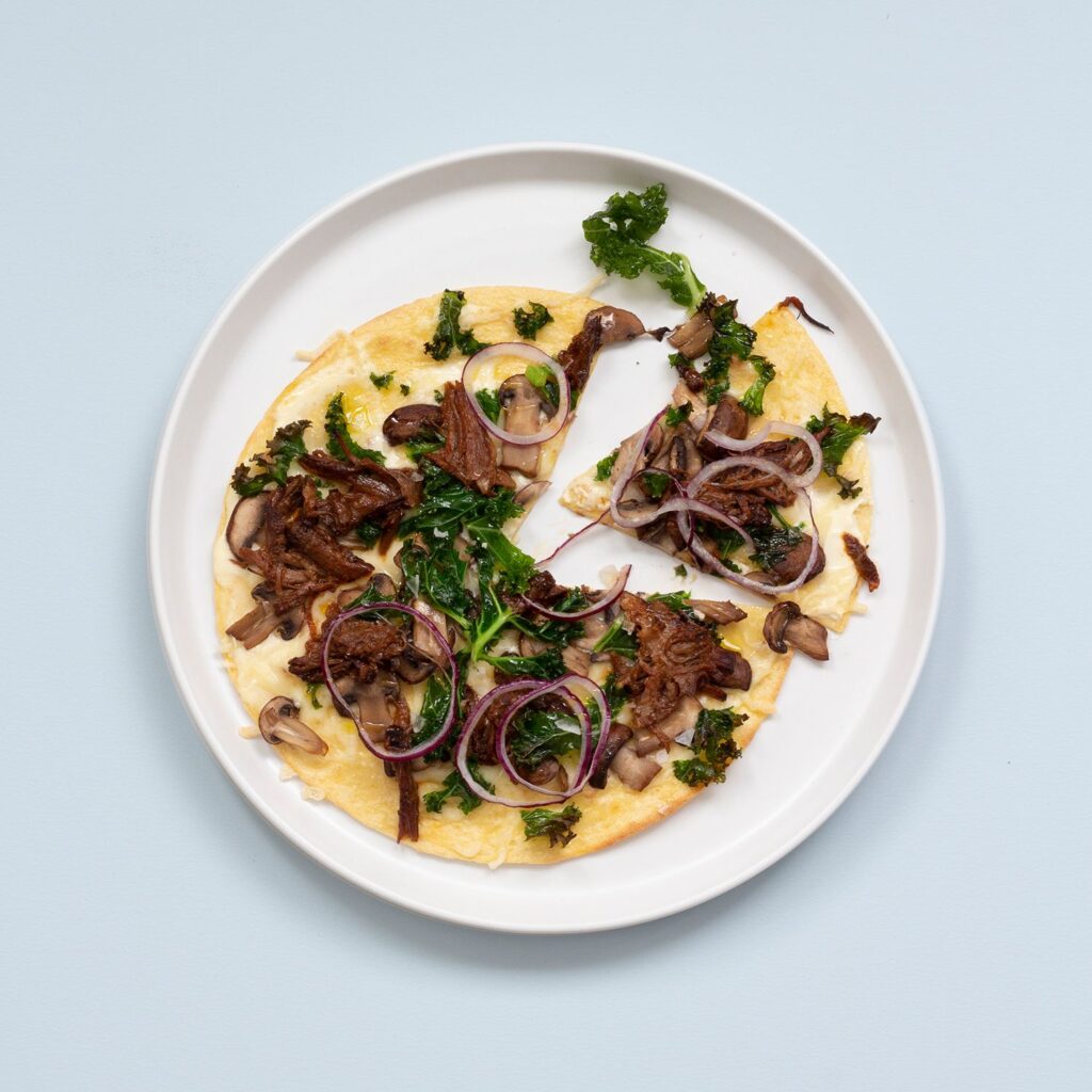 En pizza på en vit tallrik. På pizzan finns svamp, rödlök, pulled pork och grönkål
