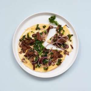 En pizza på en vit tallrik. På pizzan finns svamp, rödlök, pulled pork och grönkål