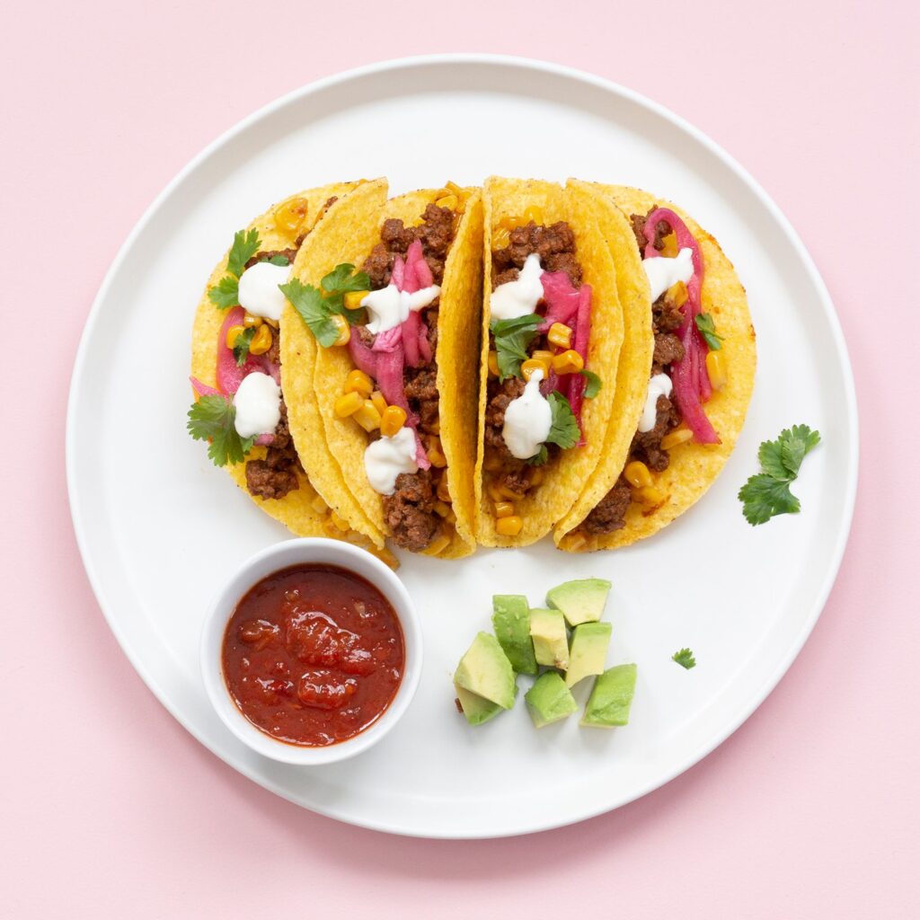 Fyra hårda tacoskal på en vit tallrik. Tacoskalen är fyllda med färs, rödlök, majs och koriander. Bredvid står en liten skål med röd salsa.