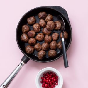 En stekpanna med köttbullar i. Bredvid står en liten skål med lingon.