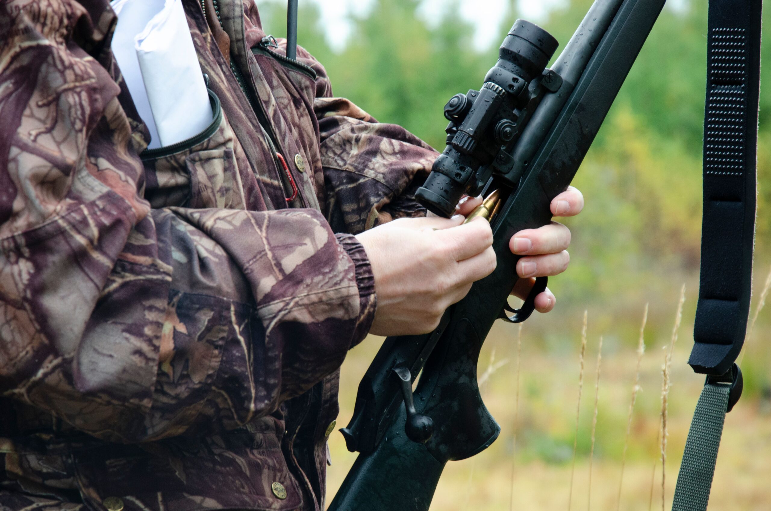 En person iförd kamouflagekläder laddar ett gevär med ett kikarsikte, stående i en naturlig skogsmiljö med suddiga gröna träd i bakgrunden.